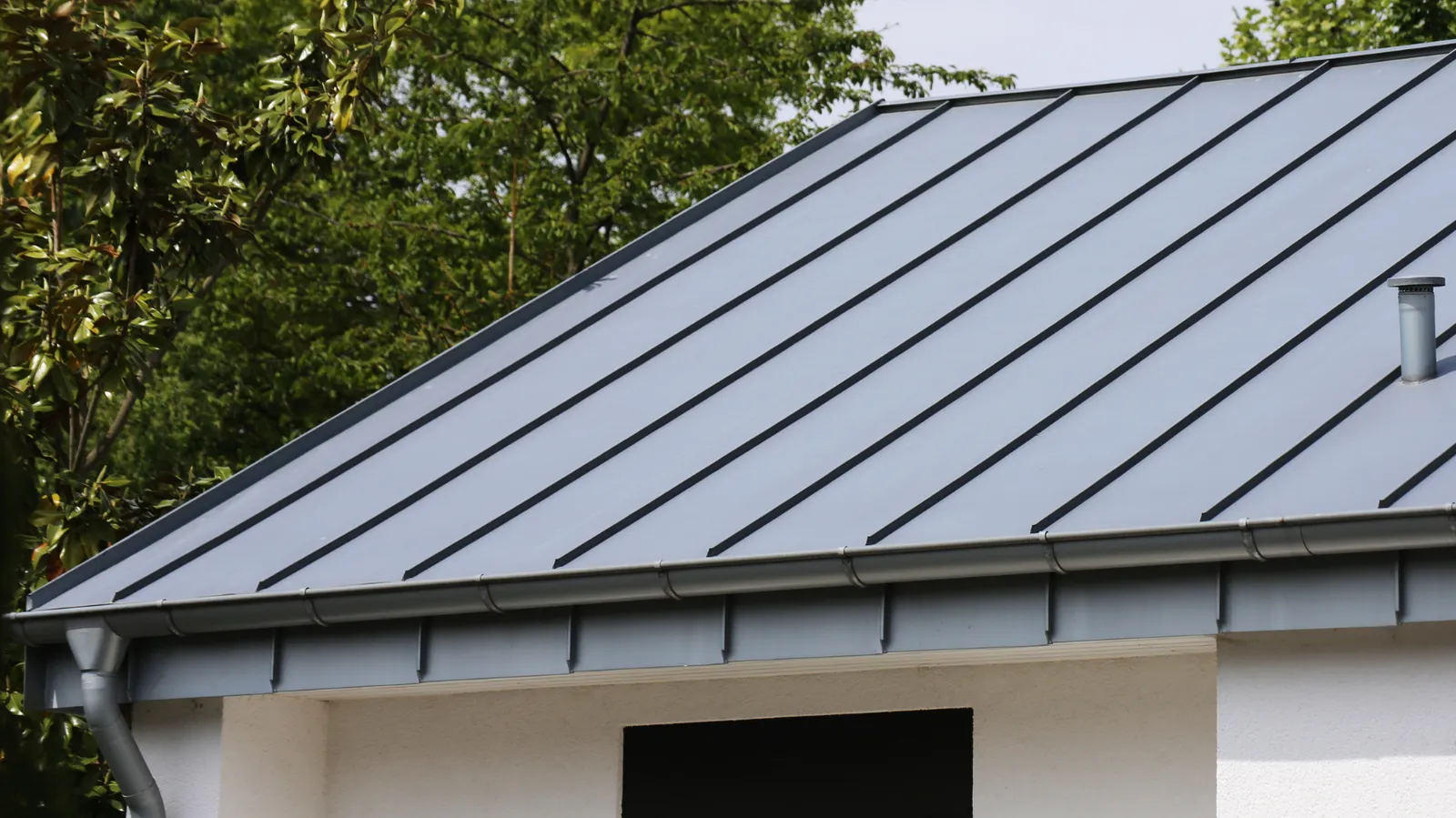 Gray standing seam metal roof with a vent pipe near trees